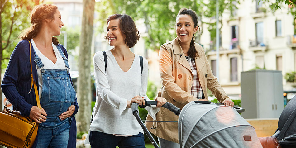 Happy pregnant woman talking to friends pushing baby strollers on sunny day